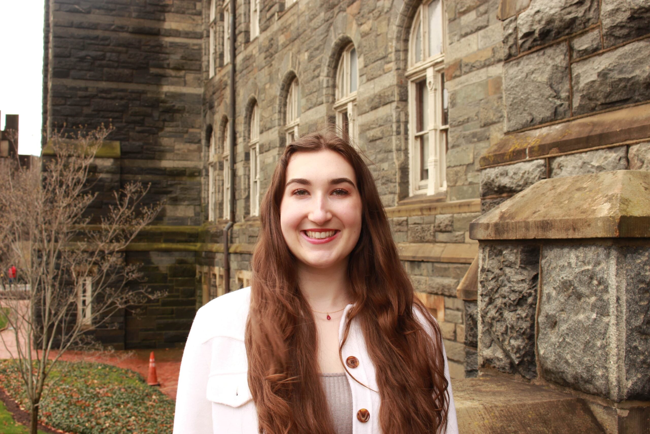 Sophia stands in front of a stone wall, wearing a white jacket and smiling brightly at the viewer. 