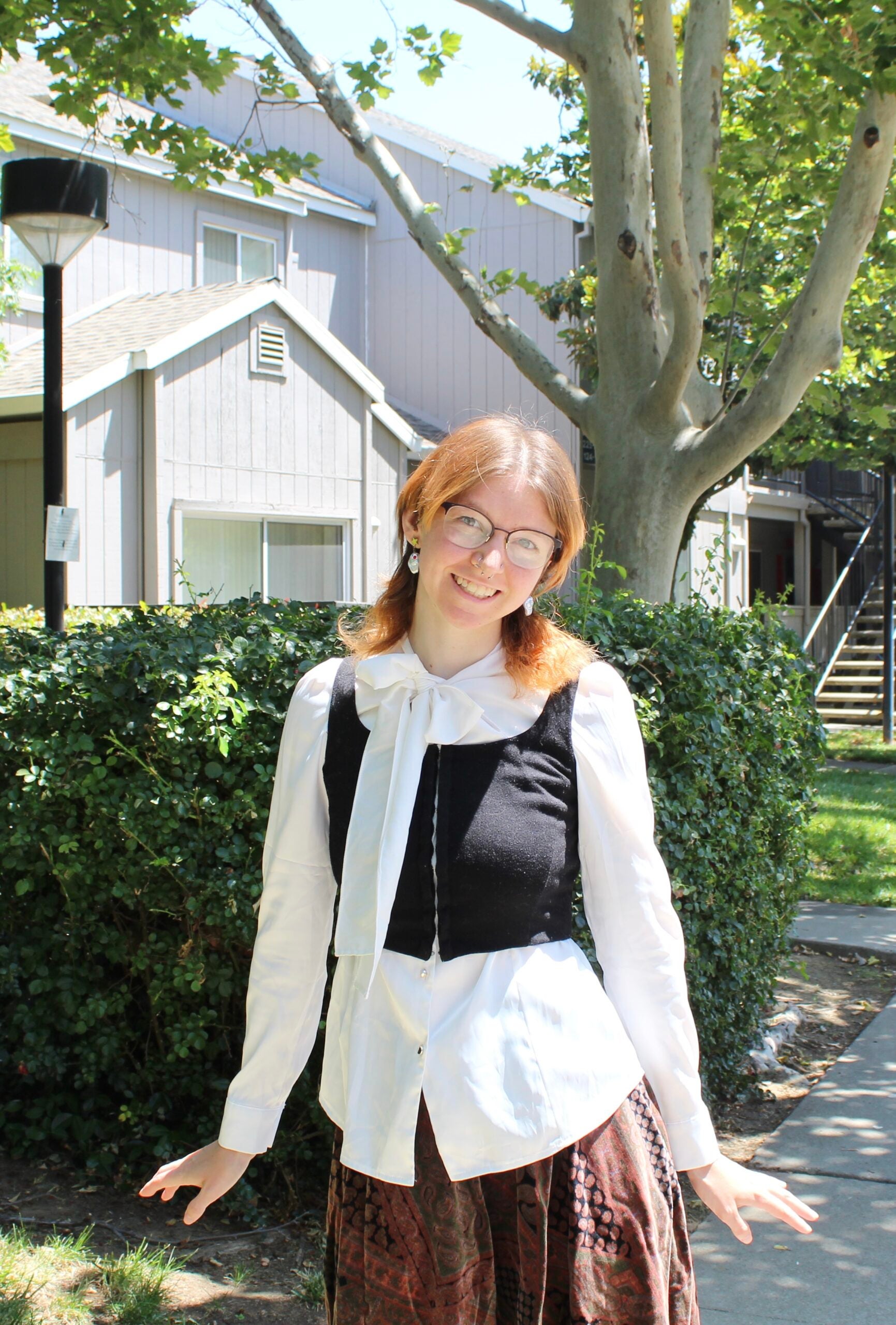 Jac stands against a suburban background, wearing a white shirt, black vest, and floral skirt, smiling at the viewer. 