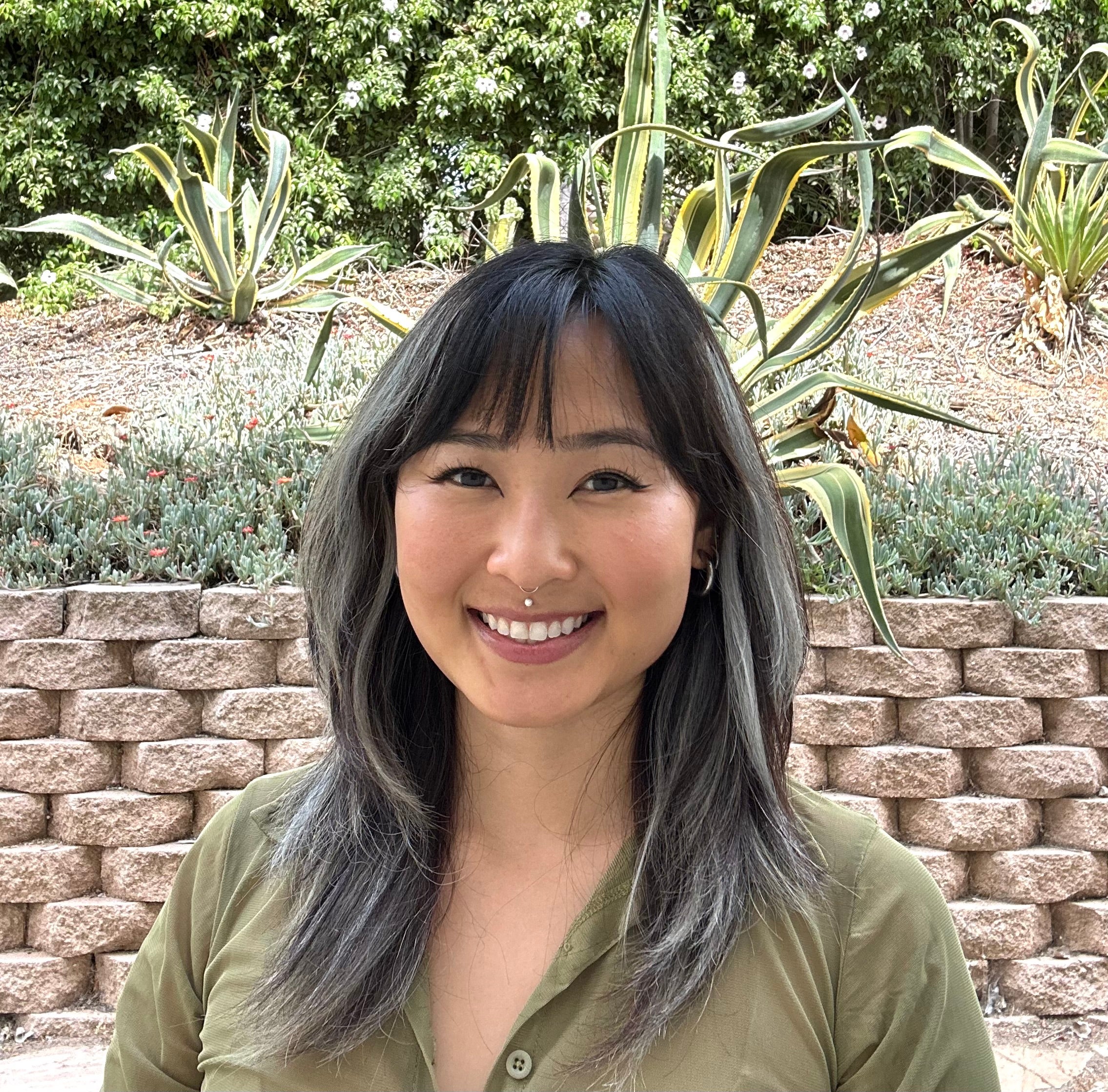 Tatiana stands in front of an adobe brick wall, wearing a green top, smiling brightly at the viewer. 