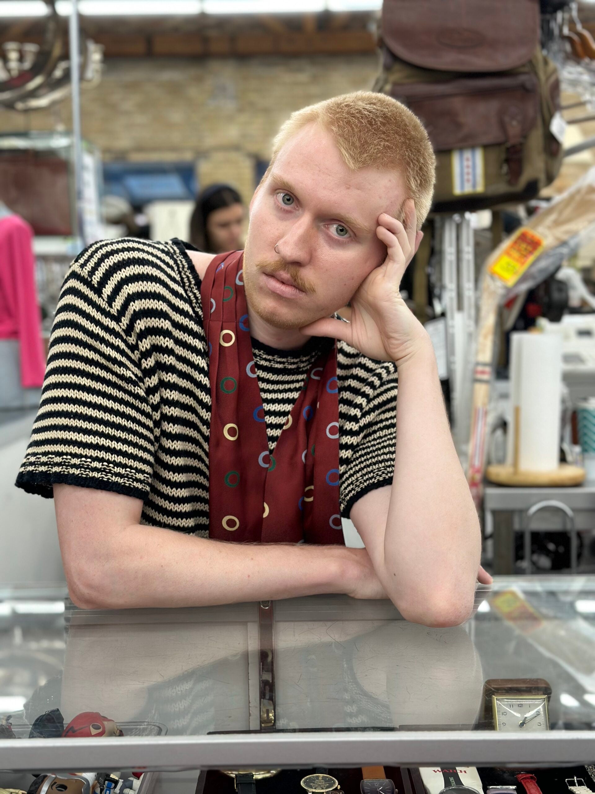 Robert leans against a glass table, wearing a striped top and scarf, looking seriously at the viewer. 
