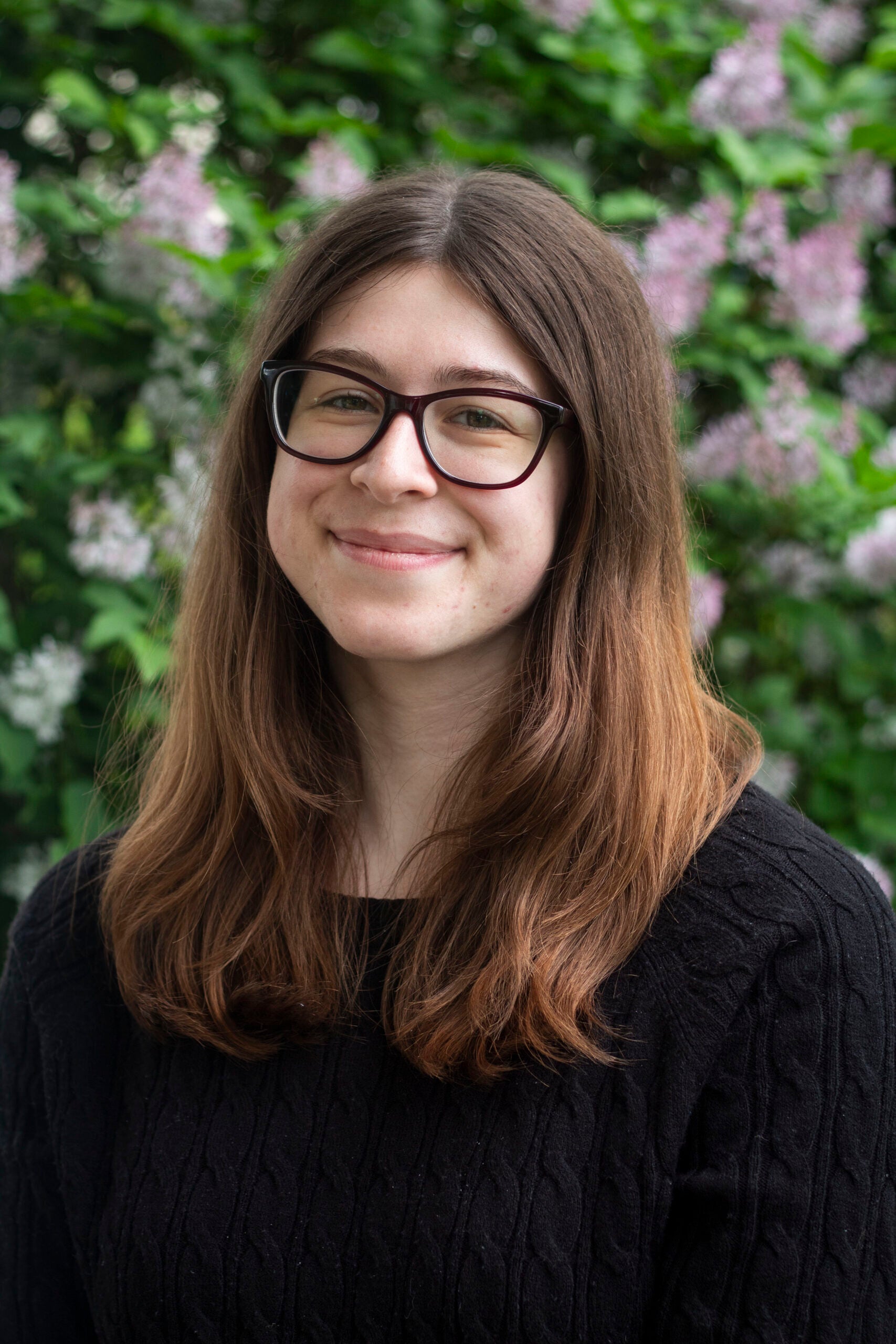Juliana Hall wearing her brown hair down over her shoulders and glasses, smiles at the camera against a flowery background. 