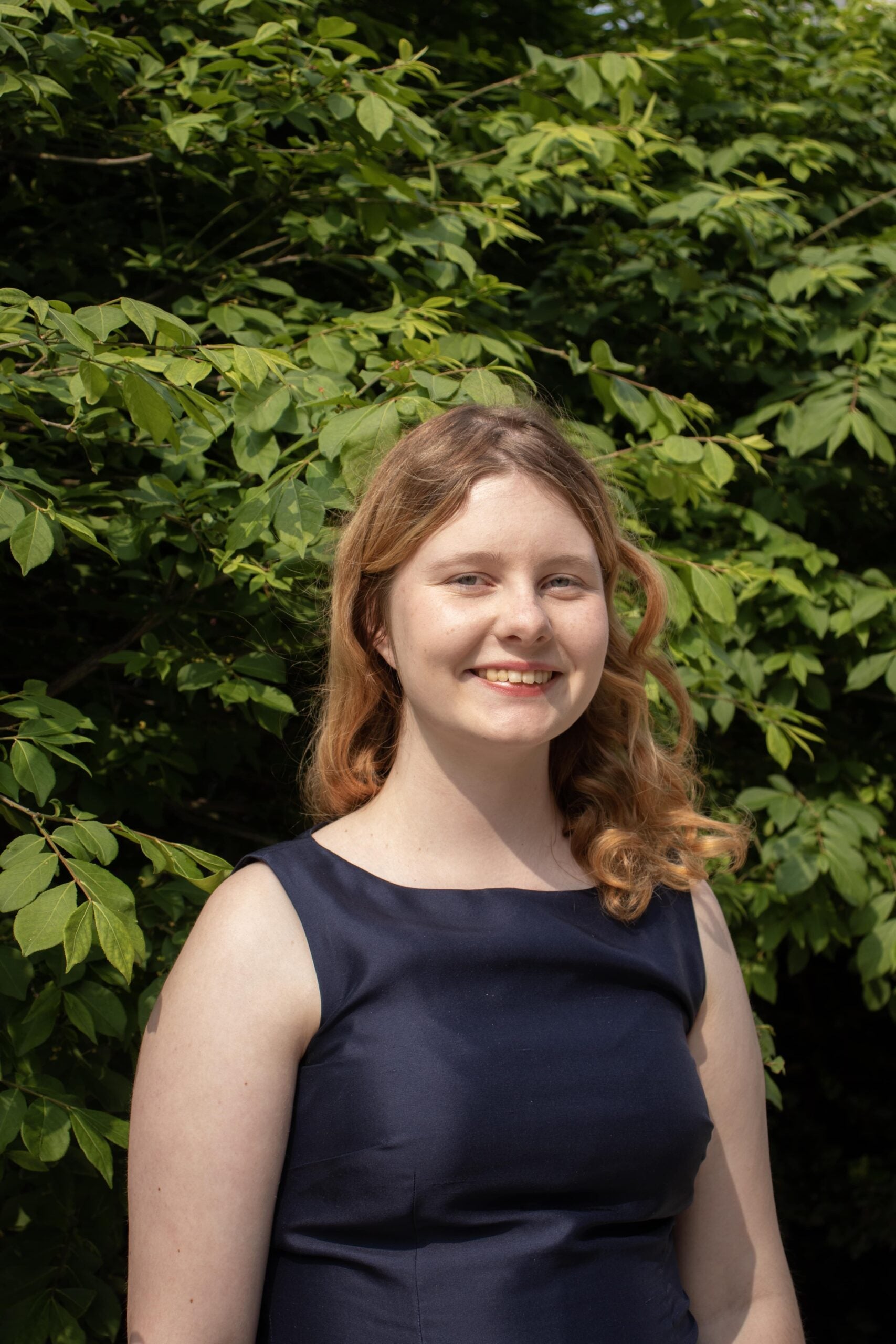 Ainsley Atwood, with blonde curls and wearing a navy blue dress, stands in front of a green background and smiles at the camera