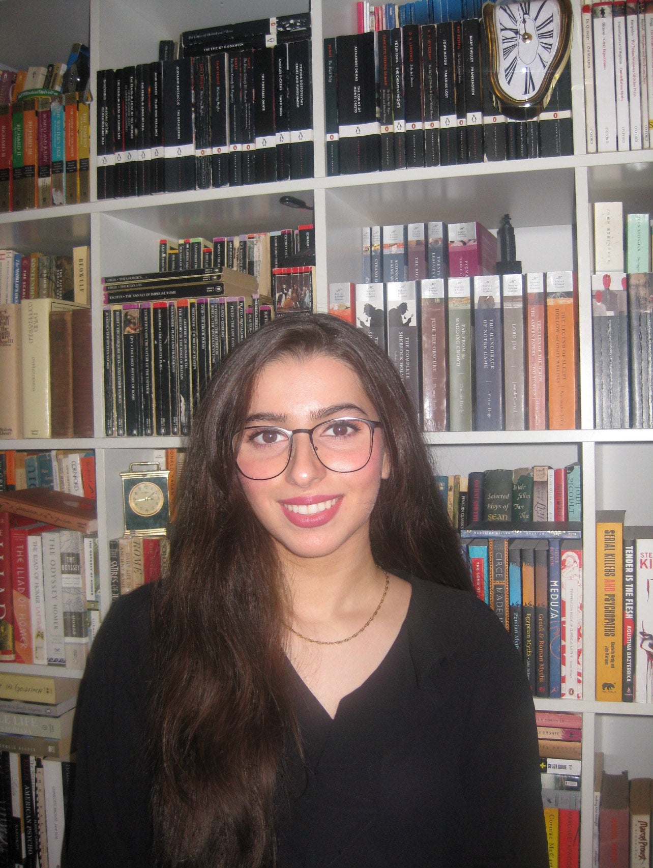 Reeba stands in front of a crowded bookshelf, wearing her dark hair down over her shoulder and a black top, smiling widely at the viewer. 