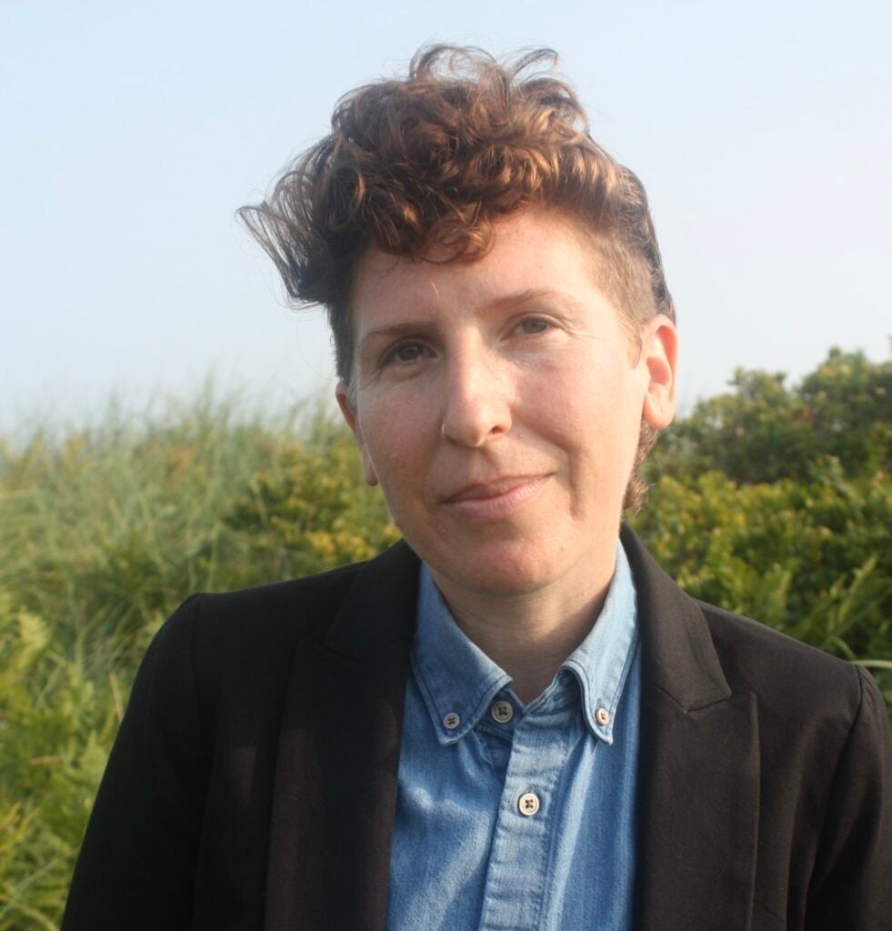 Alex Brostoff, a white person with the sides of their head shaved and a tuft of curly brown hair on top wears a jacket and blue oxford shirt in front of a green and blue background