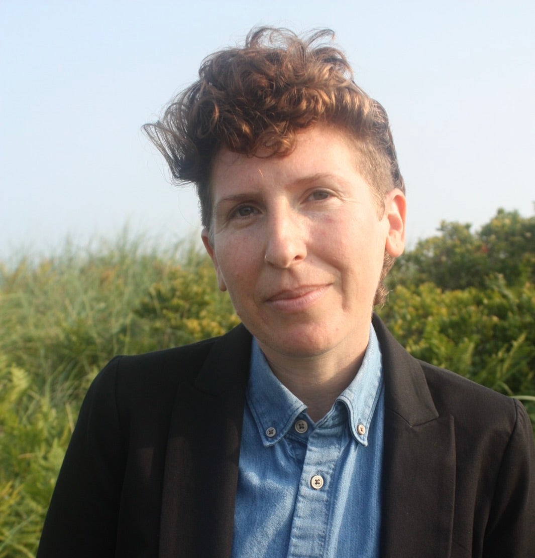 Alex Brostoff, a white person with the sides of their head shaved and a tuft of curly brown hair on top wears a jacket and blue oxford shirt in front of a green and blue background