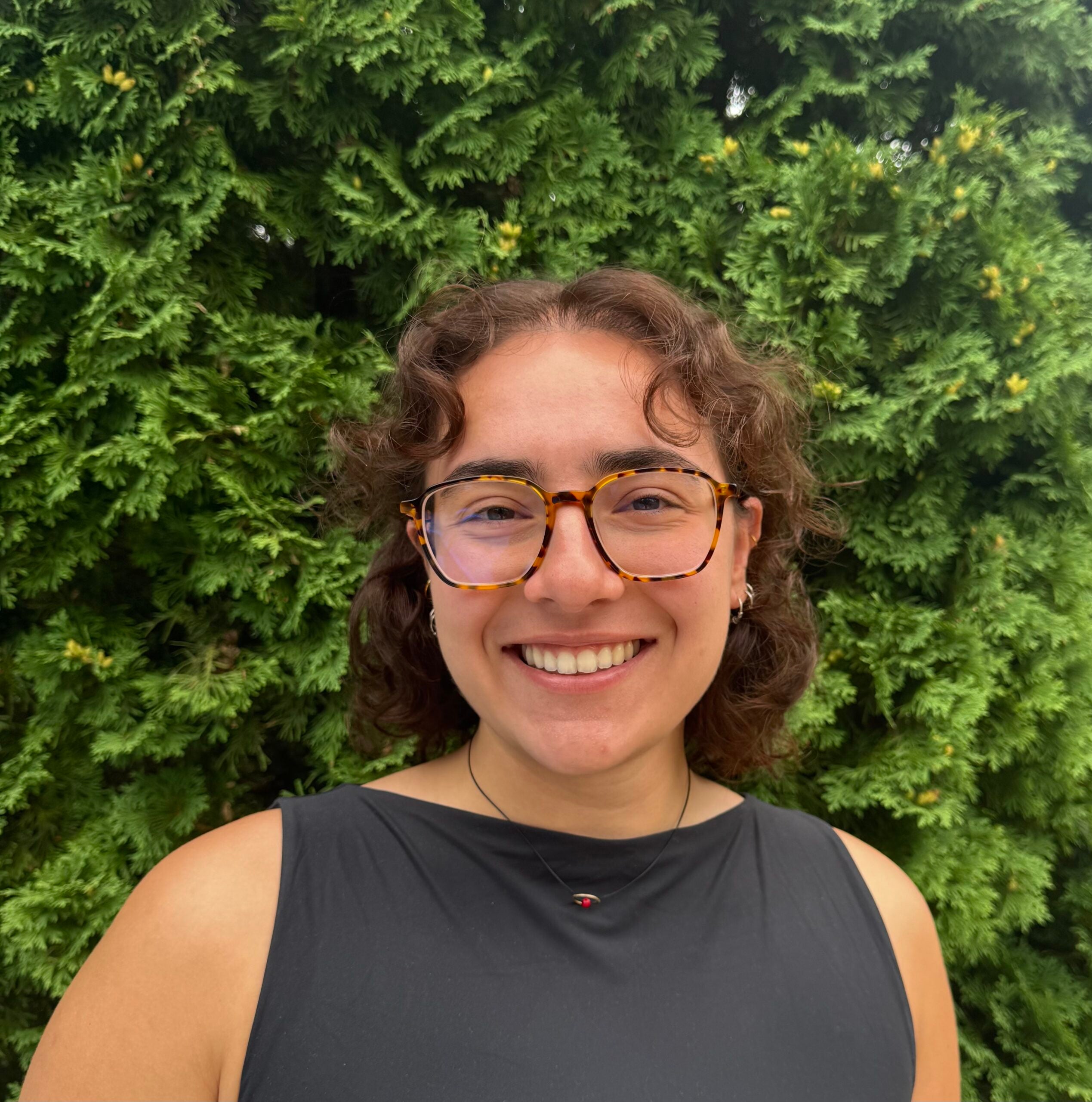 Sophia Meimaris, a young woman with curly brown hair, tortoise shell glasses and a black top smiles in front of a leafy background