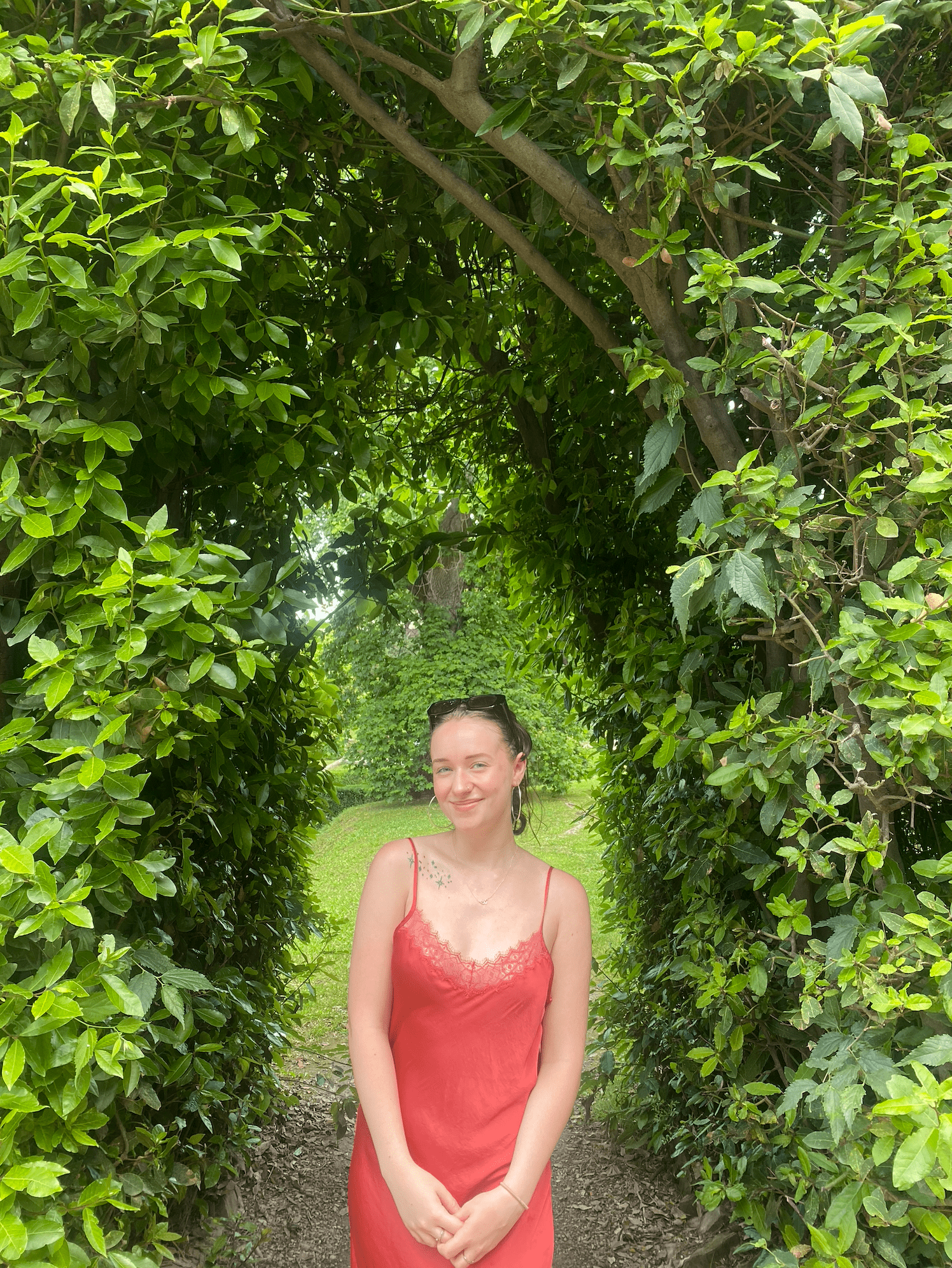 Emily Hancock stands in a green archway, wearing a pink sundress and smiling at the viewer
