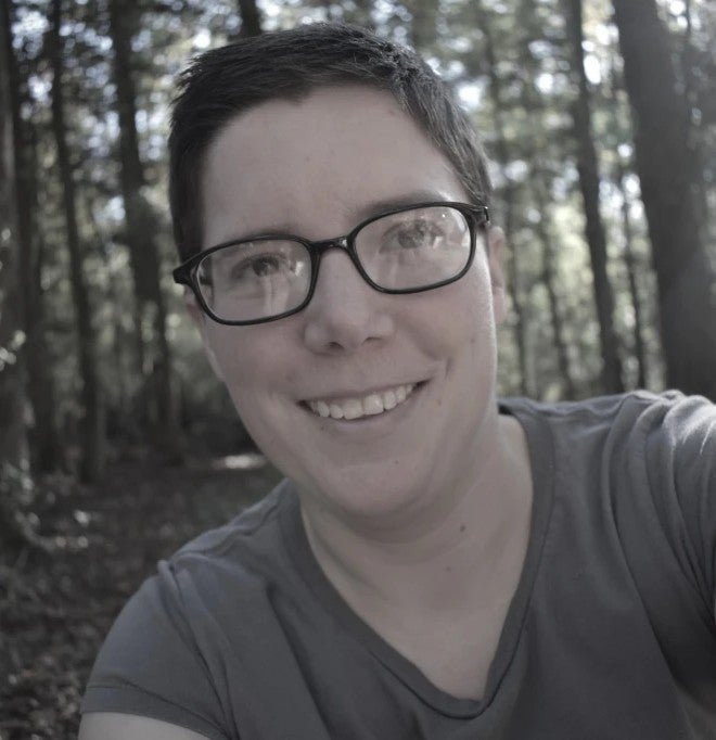 Christina, a white woman with short hair and black glasses smiles at the camera. 