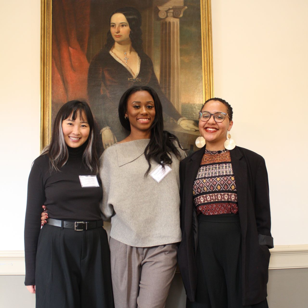 Tatiana Su, MA '27, Laurah Perrin, MA '26, and Jamira Richardson, MA '25 stand proudly in front of a portrait in the Georgetown Humanities Initiative Conference Room