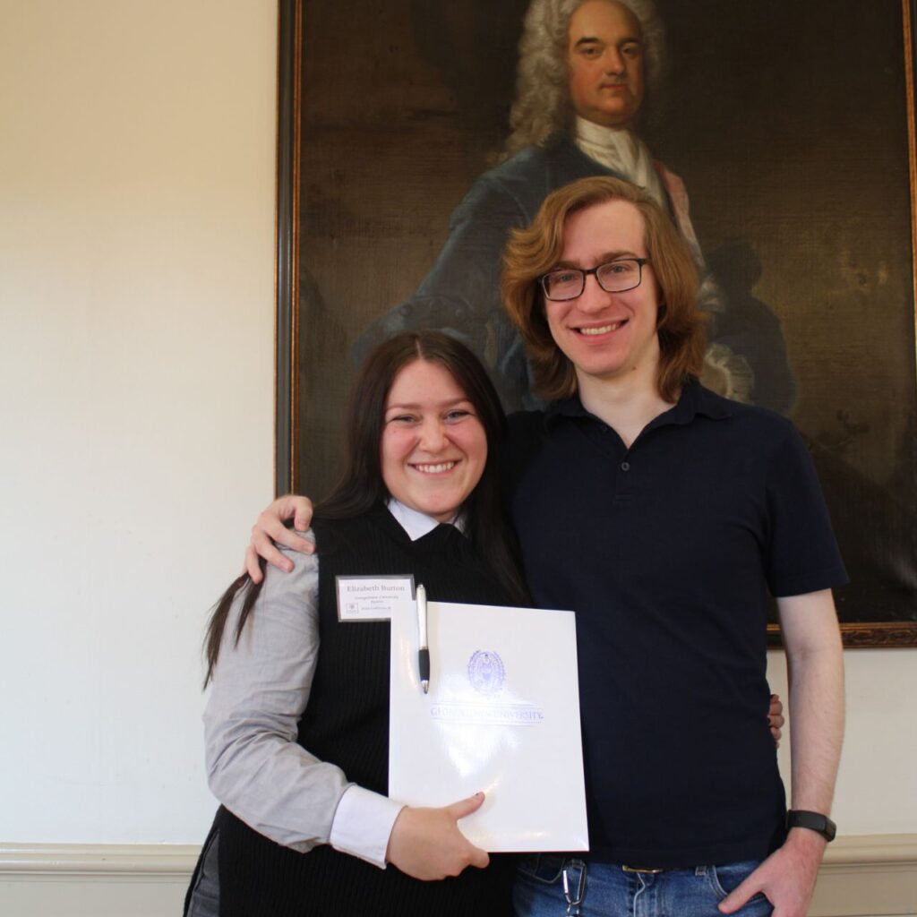 Libby Burton, MA '26, stands with her partner in front of a portrait in the Georgetown Humanities Initiative Conference Room