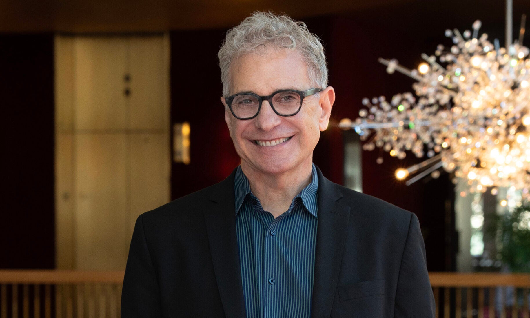 Gene Scheer, a white man in dark slacks and a dark blazer, smiles in front of a stage.