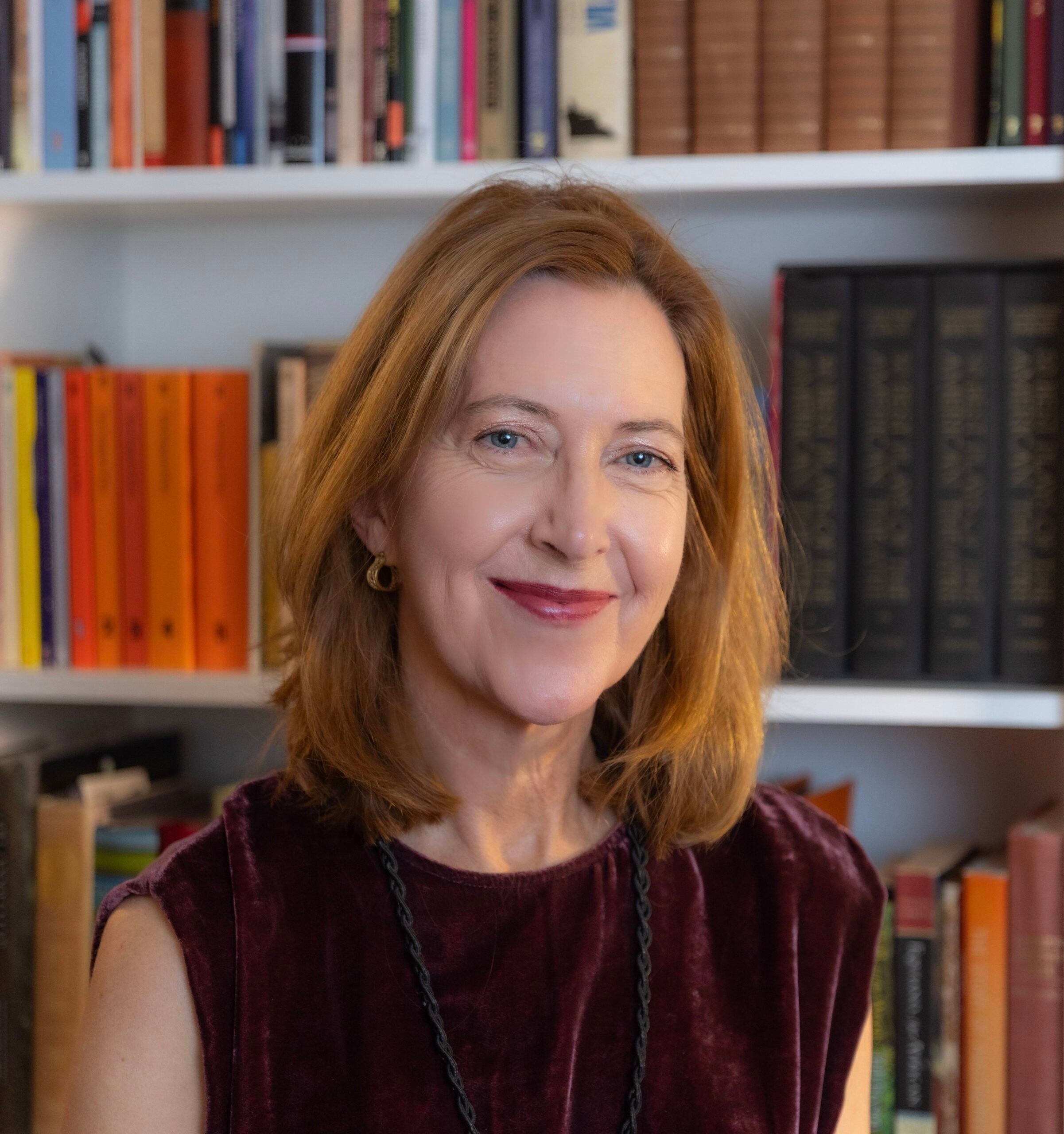 Sarah McNamer stands and smiles softly in front of a book case that is neatly appointed.