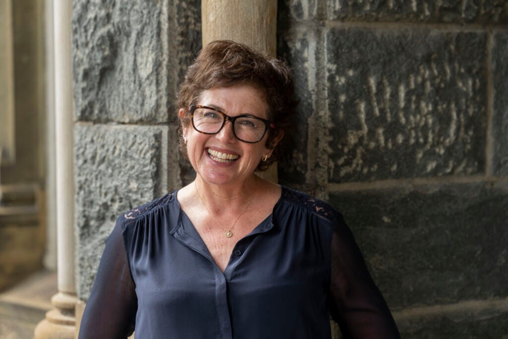 Prof. Libbie RIfkin, a white woman with short brown hair and tortoise shell glasses, smiles in a blue top while standing against a stone wall. 