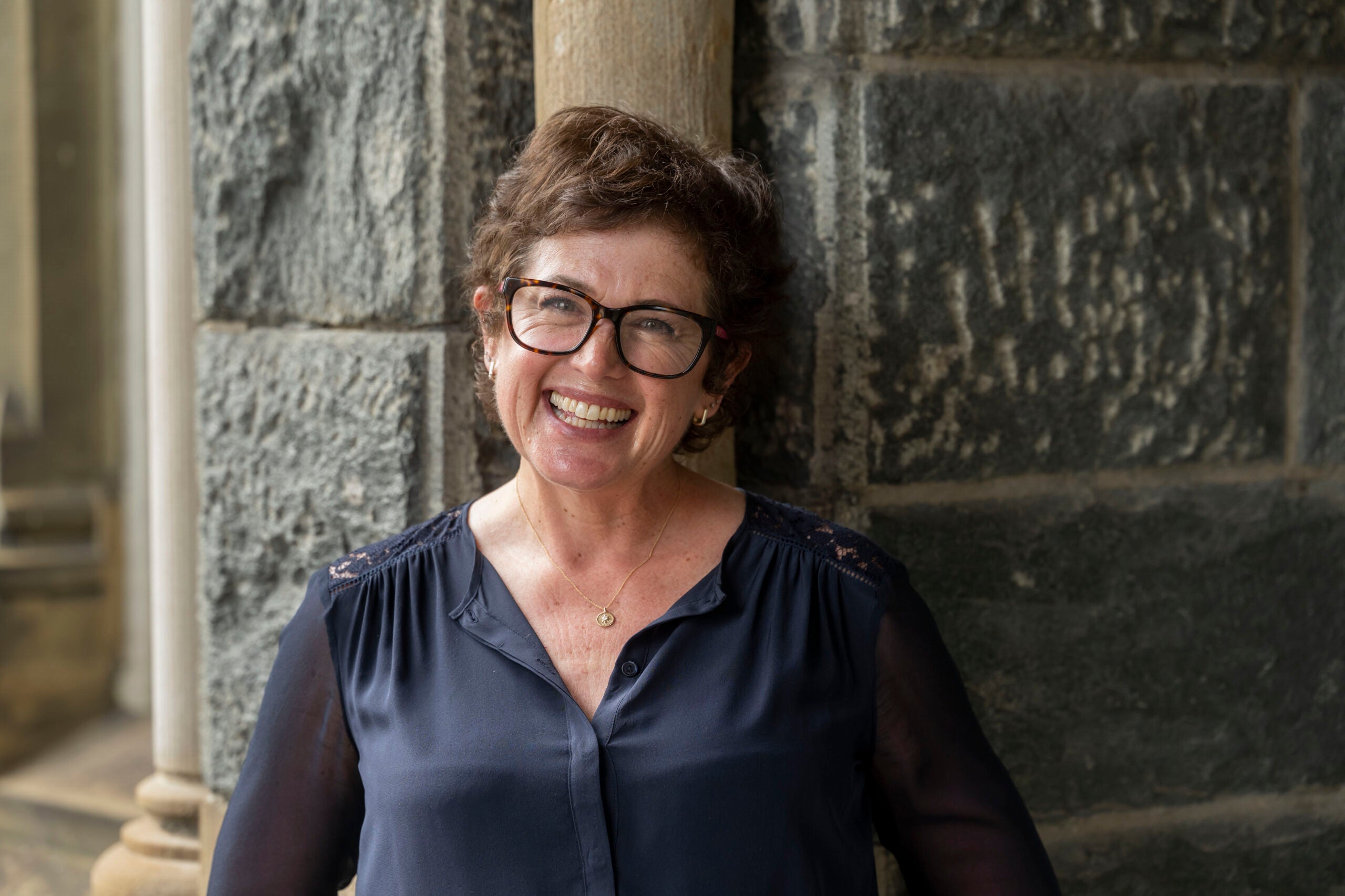 Libbie Rifkin, wearing a navy blue top and leaning against the grey stones of Healey Hall, smiles kindlyt at the viewer