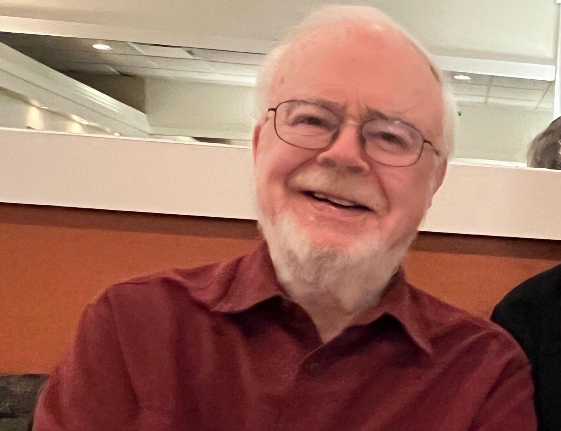 Paul Betz smiles at the camera, wearing a warm maroon flannel and leaning against a short wall